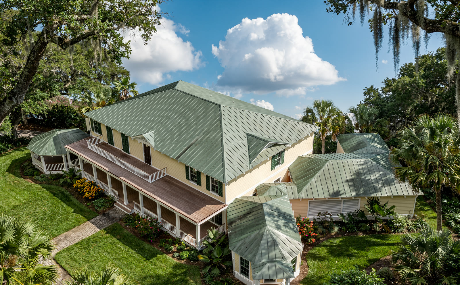 Aerial photograph of the house from above and to the
      south east. The green metal hipped roof fills the middle of the frame, the two story porch runs along the
      near face, the gazebo sits at the far left and the diagonal wing runs off to the lower right, all in a
      garden of palms and live oaks.